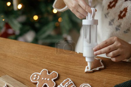 Photo for Hands decorating gingerbread cookies with icing on rustic wooden table on background of christmas golden lights. Atmospheric Christmas holiday traditions. Decorating cookies with sugar frosting - Royalty Free Image