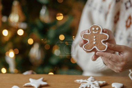 Téléchargez les photos : Joyeux Noël ! Mains tenant biscuit homme pain d'épice avec glaçage sur fond de lumières dorées arbre de Noël. Vacances de Noël dans l'atmosphère, temps de famille. - en image libre de droit