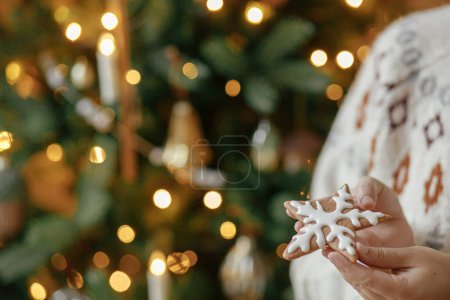 Téléchargez les photos : Joyeux Noël ! Mains tenant biscuit étoile en pain d'épice avec glaçage sur fond de lumières dorées d'arbre de Noël. Vacances de Noël dans l'atmosphère, temps de famille. - en image libre de droit
