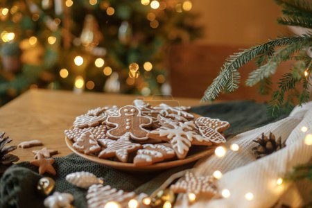 Foto de ¡Feliz Navidad! Galletas de jengibre con glaseado sobre mesa de madera con ramas de abeto y decoraciones festivas sobre fondo de luces doradas de árbol de Navidad. Víspera de Navidad atmosférica, vacaciones familiares - Imagen libre de derechos