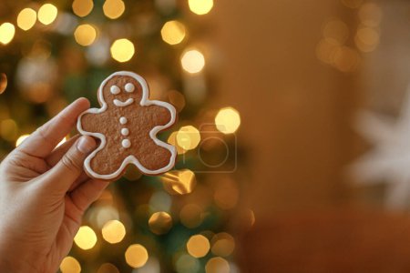 Téléchargez les photos : Main tenant pain d'épice de Noël homme cookie avec glaçage contre arbre de Noël festif avec des lumières dorées bokeh. Joyeux Noël ! Délicieux biscuits au pain d'épice, temps des fêtes atmosphériques - en image libre de droit