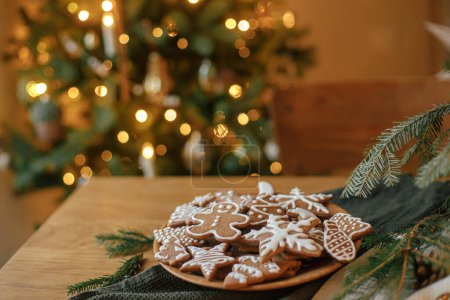 Foto de ¡Feliz Navidad! Galletas de jengibre con glaseado en plato sobre mesa de madera con ramas de abeto y decoraciones festivas contra las luces doradas de Navidad. Víspera de Navidad atmosférica, tiempo en familia - Imagen libre de derechos