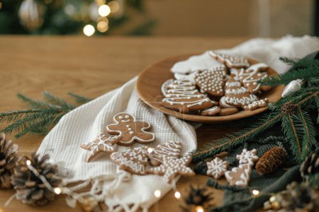 Téléchargez les photos : Joyeux Noël ! Biscuits au pain d'épice avec glaçage dans une assiette sur une table rustique en bois avec branches de sapin, décorations festives et lumières dorées de Noël. Vacances de Noël dans l'atmosphère, temps de famille - en image libre de droit
