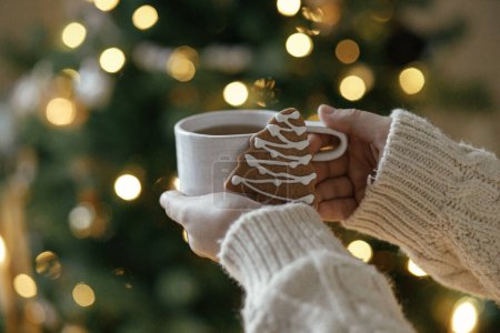 Photo for Merry Christmas! Hands in sweater holding warm cup of tea with gingerbread cookie against moody christmas tree with golden lights bokeh. Atmospheric Christmas holidays, winter hygge - Royalty Free Image