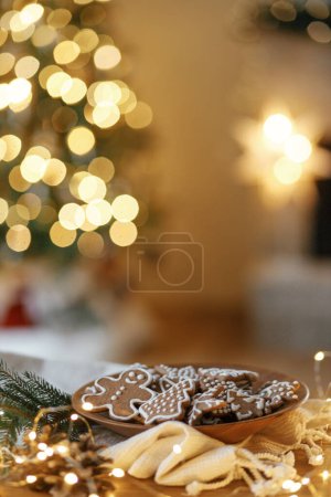 Téléchargez les photos : Biscuits au pain d'épice de Noël avec glaçage dans une assiette sur une table rustique festive avec des décorations contre l'éclairage doré. Joyeux Noël ! Délicieux biscuits au pain d'épice, veille de vacances atmosphérique - en image libre de droit