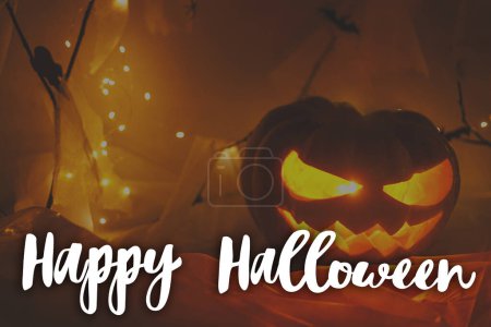 Photo for Happy Halloween! Happy Halloween text and Jack o lantern carved pumpkin with spider web and glowing lights in dark. Spooky atmospheric halloween greeting card, handwritten sign - Royalty Free Image