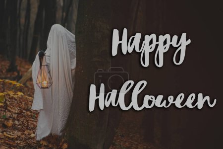 Téléchargez les photos : Joyeux Halloween ! Joyeux texte d'Halloween et fantôme effrayant tenant lanterne lumineuse dans la forêt d'automne sombre et humide. Carte de voeux halloween atmosphérique effrayante, panneau manuscrit - en image libre de droit