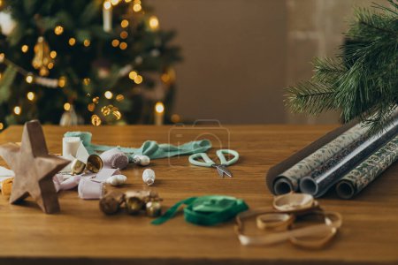 Téléchargez les photos : Emballage cadeaux de Noël. Papier d'emballage festif élégant, rubans, ciseaux, étoiles et cloches en laiton sur une table en bois sur fond de lumières d'arbre de Noël. Préparation des vacances d'hiver - en image libre de droit