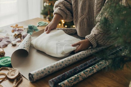 Foto de Envolver regalos de Navidad. Manos en suéter acogedor que envuelve suéter elegante en papel de regalo festivo con cintas, adornos vintage, arcos en la mesa de madera. Vacaciones de invierno atmosféricas - Imagen libre de derechos