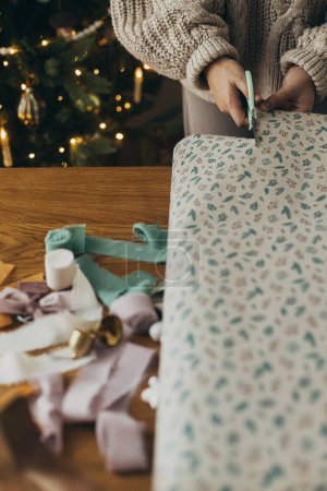 Téléchargez les photos : Emballage cadeaux de Noël. Mains dans le pull confortable tenant élégant papier d'emballage festif avec des rubans, des ornements vintage, des arcs sur la table en bois. Vacances d'hiver dans l'atmosphère - en image libre de droit