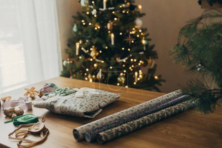 Téléchargez les photos : Emballage cadeaux de Noël. Élégant cadeau, papier d'emballage festif, rubans, ciseaux, étoiles et cloches en laiton sur une table en bois sur fond de lumières de sapin de Noël. Préparation des vacances d'hiver - en image libre de droit