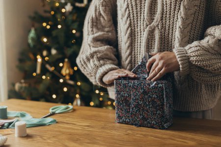 Téléchargez les photos : Emballage cadeaux de Noël. Mains dans un pull confortable enveloppant cadeau élégant dans du papier d'emballage festif avec des rubans, des ornements vintage, des arcs sur une table en bois. Vacances d'hiver dans l'atmosphère - en image libre de droit