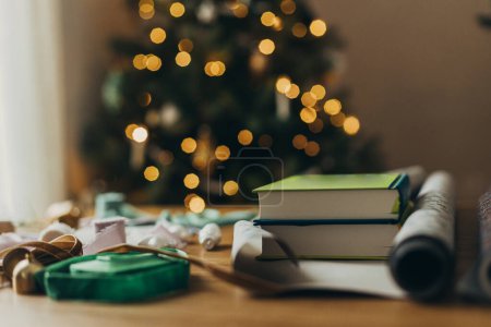 Téléchargez les photos : Emballage cadeaux de Noël. Livres élégants comme présents en papier d'emballage festif, rubans, ornements vintage sur table en bois sur fond de lumières d'arbre de Noël. Vacances d'hiver dans l'atmosphère - en image libre de droit