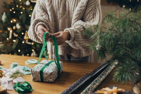Foto de Mujer en suéter acogedor envolviendo elegante regalo de Navidad en papel de regalo festivo con cintas, adornos vintage, arcos en la mesa de madera. Vacaciones de invierno atmosféricas, envolviendo regalo de Navidad - Imagen libre de derechos