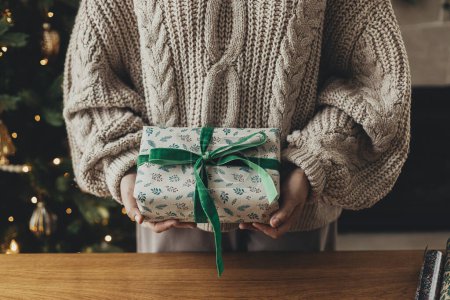 Téléchargez les photos : Joyeux Noël ! Femme en pull confortable tenant cadeau de Noël élégant en papier d'emballage festif avec arc, ruban sur table en bois. Vacances d'hiver dans l'atmosphère - en image libre de droit