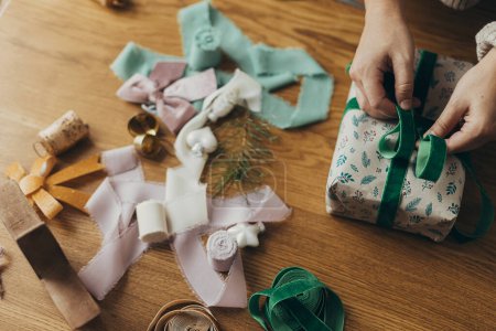 Téléchargez les photos : Femme en pull confortable enveloppant cadeau de Noël élégant en papier d'emballage festif avec des rubans, des ornements vintage, des arcs sur la table en bois vue sur le dessus. Vacances dans l'atmosphère, emballage cadeau de Noël - en image libre de droit