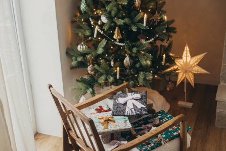 Téléchargez les photos : Cadeaux de Noël élégants en papier d'emballage festif avec des arcs, des ornements vintage, un jouet sur chaise sur fond d'étoile de Noël dorée et des lumières d'arbre. Joyeuses fêtes ! Veille de Noël atmosphérique - en image libre de droit