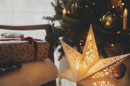 Téléchargez les photos : Élégantes étoiles de Noël dorées et cadeaux de Noël en papier d'emballage festif avec arcs, ornements vintage sur chaise sous les lumières des arbres. Joyeuses fêtes ! Veille de Noël atmosphérique - en image libre de droit