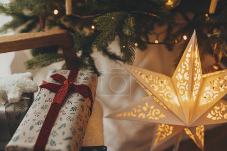 Téléchargez les photos : Cadeaux de Noël élégants en papier d'emballage festif avec des arcs, des ornements vintage sur la chaise sur fond d'étoile de Noël dorée et des lumières d'arbre. Joyeuses fêtes ! Veille de Noël atmosphérique - en image libre de droit