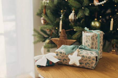 Foto de ¡Feliz Navidad! Elegantes regalos de Navidad envueltos en papel festivo con lazos, cintas vintage, juguetes en la mesa de madera contra las luces del árbol de Navidad. Vacaciones de invierno atmosféricas - Imagen libre de derechos