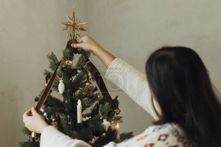 Photo for Decorating christmas tree with stylish golden star and ribbon. Woman in cozy sweater hanging vintage ornament on tree branch close up. Atmospheric winter holiday tradition, family time - Royalty Free Image