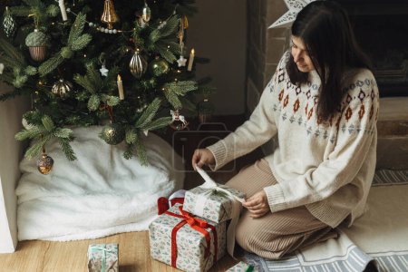 Photo for Woman in cozy sweater wrapping stylish christmas gifts under decorated christmas tree with modern vintage baubles. Atmospheric winter holidays time. Preparing Christmas presents - Royalty Free Image