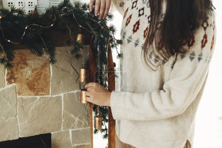 Téléchargez les photos : Décorer cheminée de Noël avec ruban de velours et cloches vintage sur branches de sapin. Femme en pull confortable accrochant une décoration élégante sur le manteau dans une ferme moderne. Vacances d'hiver dans l'atmosphère - en image libre de droit