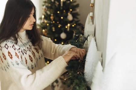 Téléchargez les photos : Femme en pull confortable accrochant une décoration élégante sur le manteau dans une ferme moderne. Décorer cheminée de Noël avec des lumières et des cloches vintage sur les branches de sapin. Vacances d'hiver dans l'atmosphère - en image libre de droit