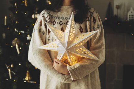 Téléchargez les photos : Joyeux Noël ! Femme en pull confortable tenant illuminé étoile de Noël sur fond d'arbre de Noël décoré avec des boules et des lumières vintage. Veille de Noël atmosphérique - en image libre de droit