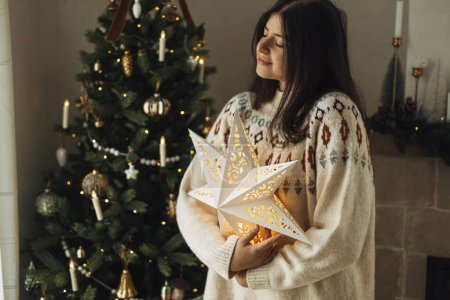 Foto de ¡Feliz Navidad! Mujer en suéter acogedor celebración iluminada estrella de Navidad en el fondo del árbol de Navidad decorado con adornos vintage y luces. Nochebuena atmosférica - Imagen libre de derechos