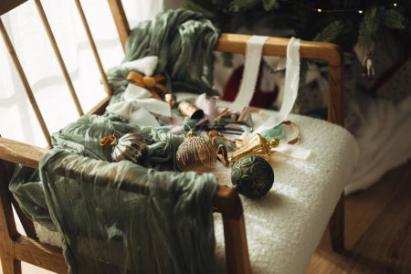 Téléchargez les photos : Élégantes boules vintage de Noël, rubans, cloches en laiton et ciseaux sur une chaise blanche confortable sous l'arbre. Ornements de Noël esthétique. Décorer l'arbre de Noël. Joyeux Noël! - en image libre de droit