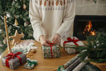 Foto de Mujer envolviendo elegante regalo de Navidad con cinta roja en la mesa de madera con decoraciones festivas en la habitación escandinava decorada. Feliz Navidad y Felices Fiestas! Presente de la mano - Imagen libre de derechos