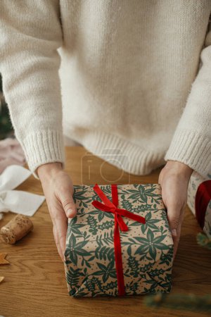 Foto de Mujer sosteniendo elegantes regalos de Navidad con cinta roja en la mesa de madera con decoraciones festivas en la habitación escandinava. Feliz Navidad y felices fiestas! - Imagen libre de derechos