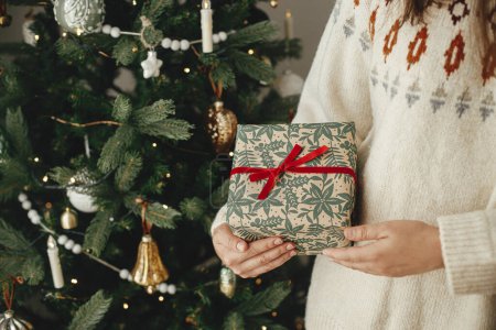 Foto de Feliz Navidad y felices fiestas! Mujer sosteniendo elegante regalo de Navidad con cinta de cerca en el fondo del árbol decorado moderno en la habitación escandinava. - Imagen libre de derechos