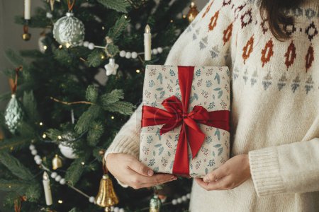 Téléchargez les photos : Joyeux Noël et Joyeuses fêtes ! Femme en pull tenant élégant cadeau de Noël avec ruban rouge à l'arbre vintage décoré moderne dans la chambre scandinave. - en image libre de droit