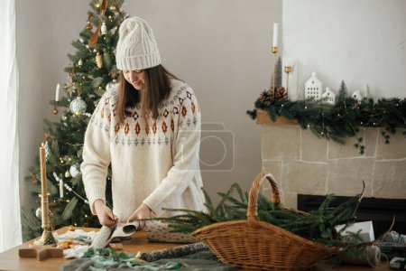 Photo for Woman in cozy sweater and hat wrapping christmas gift in paper on wooden table with festive decorations in decorated scandinavian room. Merry Christmas! Hands packing present - Royalty Free Image