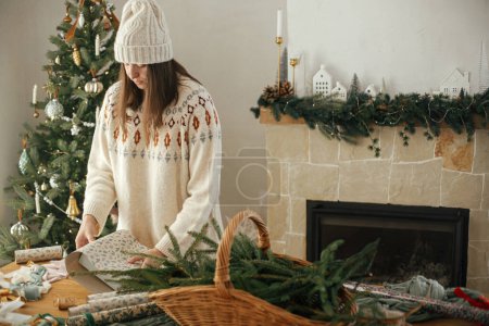Photo for Woman in cozy sweater and hat wrapping christmas gift in paper on wooden table with festive decorations in decorated scandinavian room. Merry Christmas! Hands packing present - Royalty Free Image
