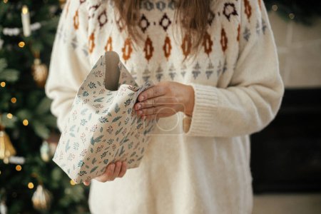Foto de Mujer en suéter acogedor envolviendo elegante regalo de Navidad en papel sobre el fondo del elegante árbol de Navidad en la habitación escandinava decorativa. ¡Feliz Navidad! Manos regalo de embalaje - Imagen libre de derechos