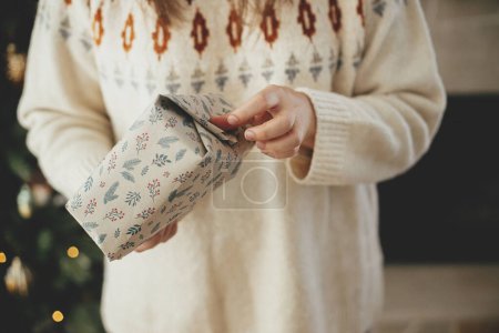 Foto de Mujer en suéter acogedor envolviendo elegante regalo de Navidad en papel sobre el fondo del elegante árbol de Navidad en la habitación escandinava decorativa. ¡Feliz Navidad! Manos regalo de embalaje - Imagen libre de derechos