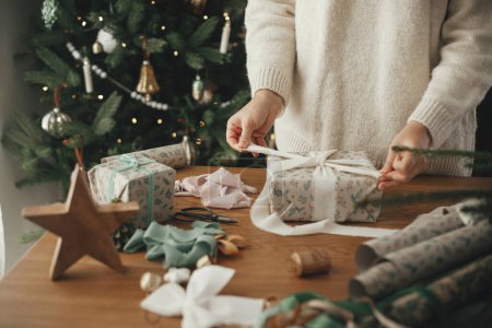 Téléchargez les photos : Femme en pull enveloppant cadeau de Noël avec ruban élégant sur une table en bois avec des décorations festives dans une chambre scandinave décorée. Joyeux Noël ! Emballage des mains présent gros plan - en image libre de droit