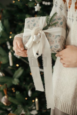 Téléchargez les photos : Femme tenant cadeau de Noël élégant avec ruban et branche de sapin près sur le fond de l'arbre décoré moderne dans la chambre scandinave. Joyeux Noël et joyeuses fêtes! - en image libre de droit