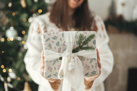 Téléchargez les photos : Joyeux Noël et bonnes vacances ! Femme tenant cadeau de Noël élégant avec ruban et branche de sapin gros plan sur fond d'arbre décoré moderne dans la chambre scandinave. - en image libre de droit