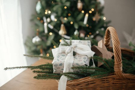 Téléchargez les photos : Joyeux Noël et Joyeuses fêtes ! Élégant cadeau de Noël enveloppé, panier rustique avec branches de sapin et décorations modernes sur table en bois contre arbre décoré festif dans la chambre scandinave - en image libre de droit