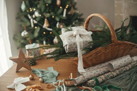 Foto de Elegante regalo de Navidad envuelto, cesta rústica con ramas de abeto y decoraciones modernas en mesa de madera contra el árbol decorado festivo en la habitación escandinava. Feliz Navidad y Felices Fiestas! - Imagen libre de derechos