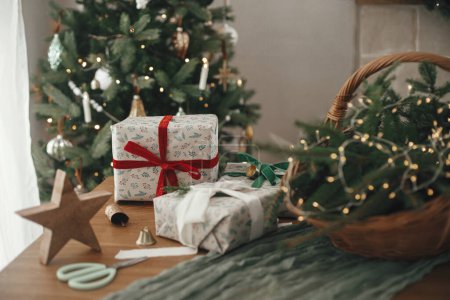 Téléchargez les photos : Cadeaux de Noël élégants enveloppés, panier rustique avec branches de sapin et décorations modernes contre arbre décoré festif dans la chambre scandinave. Joyeux Noël et Joyeuses fêtes! - en image libre de droit