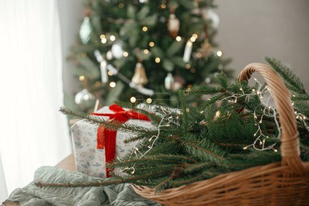 Foto de Elegante cesta rústica con ramas de abeto, regalos de Navidad envueltos y decoraciones modernas contra el árbol decorado festivo en la habitación escandinava. Feliz Navidad y Felices Fiestas! - Imagen libre de derechos