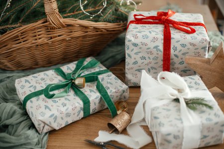 Foto de Feliz Navidad y Felices Fiestas! Elegantes regalos de Navidad envueltos, cesta rústica con ramas de abeto y decoraciones modernas en la mesa en la habitación escandinava decorada festiva - Imagen libre de derechos