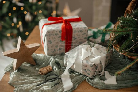 Foto de Feliz Navidad y Felices Fiestas! Elegantes regalos de Navidad envueltos, cesta rústica con ramas de abeto y decoraciones modernas en la mesa en la habitación escandinava decorada festiva. Imagen atmosférica - Imagen libre de derechos