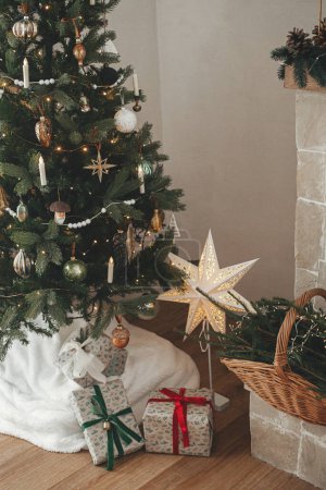 Téléchargez les photos : Cadeaux de Noël élégants enveloppés et panier rustique avec branches de sapin sous arbre de Noël décoré festive dans la chambre scandinave. Joyeux Noël et Joyeuses fêtes! - en image libre de droit