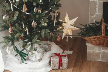 Téléchargez les photos : Cadeaux de Noël élégants enveloppés et panier rustique avec branches de sapin sous arbre de Noël décoré festive dans la chambre scandinave. Joyeux Noël et Joyeuses fêtes! - en image libre de droit
